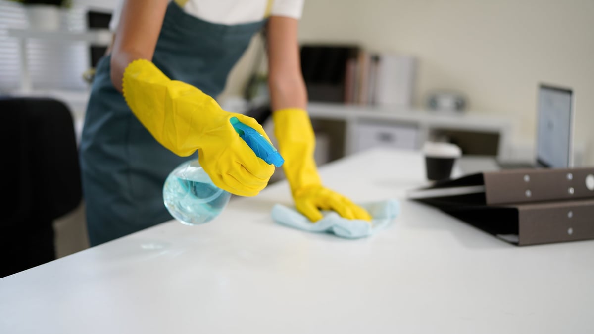Cleaner wearing yellow gloves wiping a white table with disinfectant spray