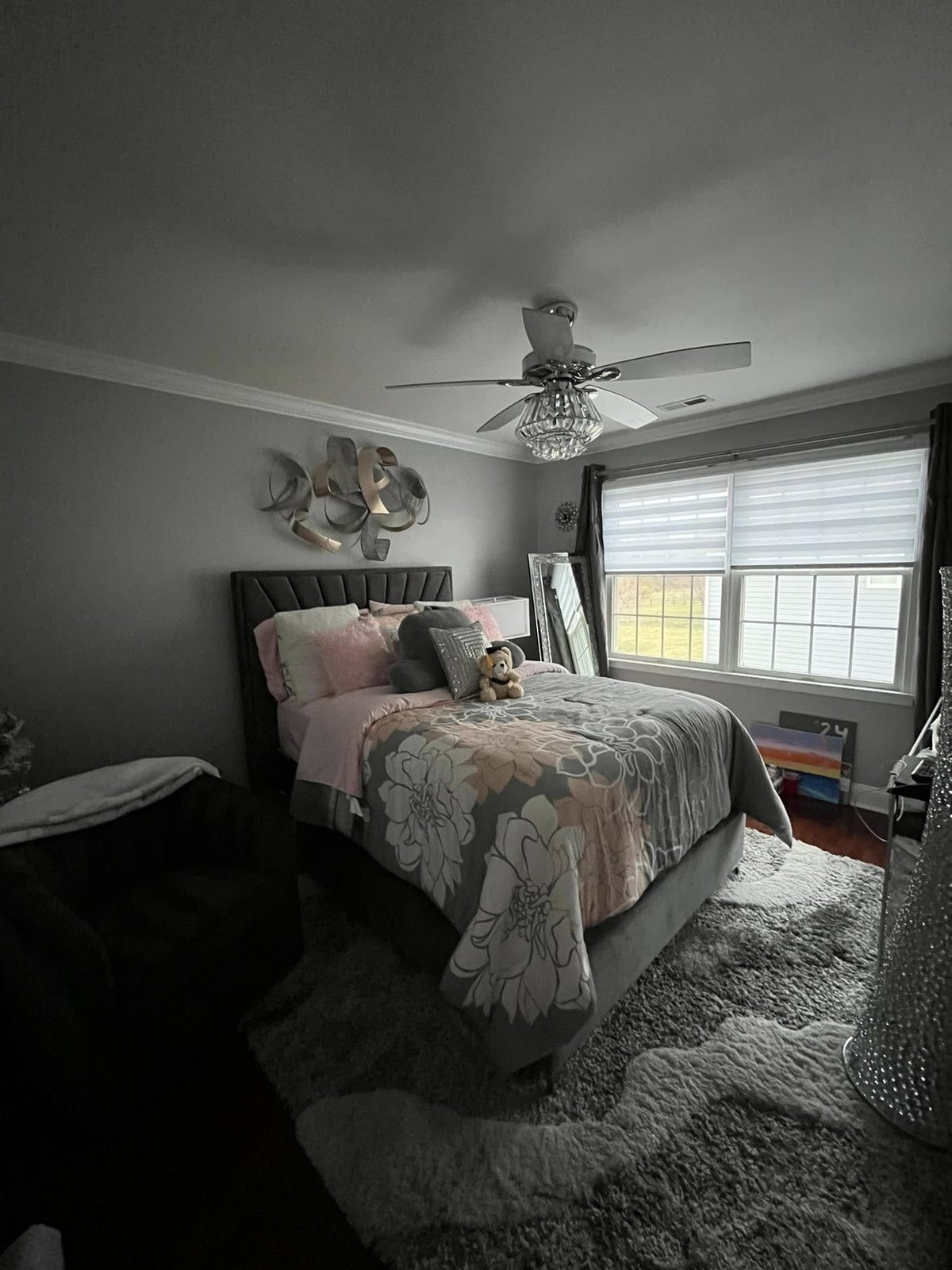 Modern bedroom with gray bed, ceiling fan, wall art, and windows with white blinds overlooking a residential area