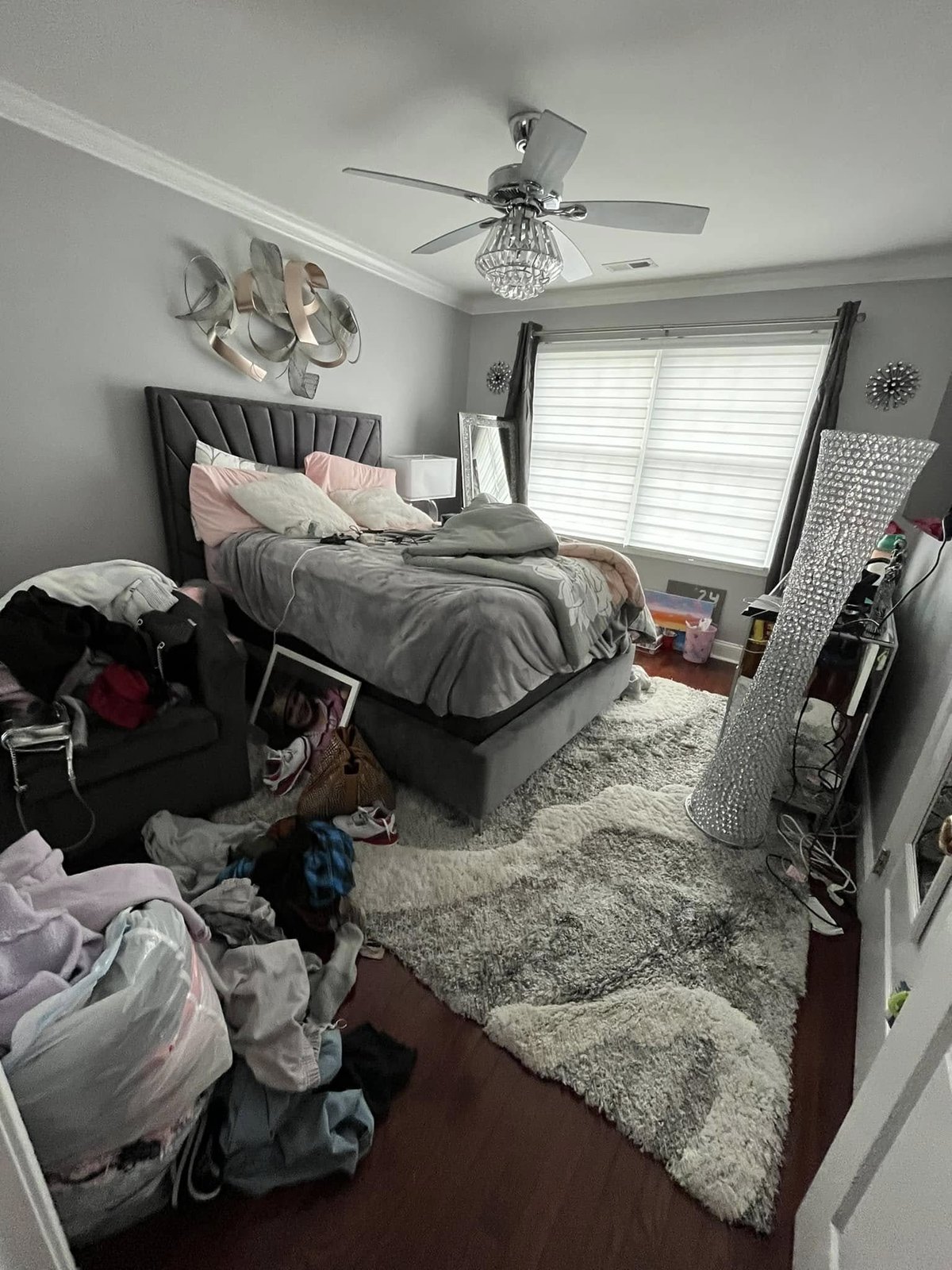 Bedroom with unmade bed, ceiling fan, large windows, cluttered floor with clothes and items scattered around